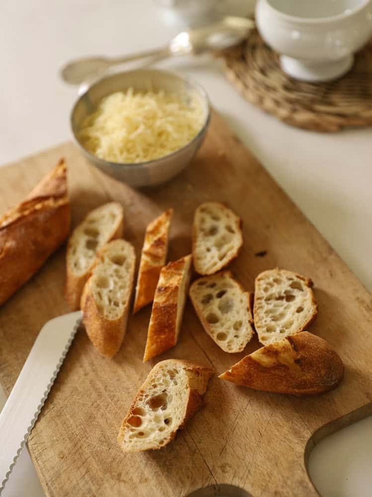 sliced day old baguette, bowls and grated Gruyere cheese on a cutting board.
