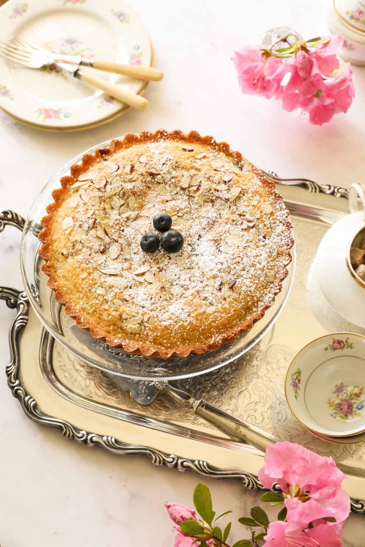 a blueberry almond cake on a glass cake stand on a tray with tea cups