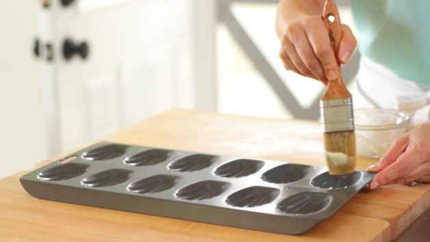 Madeleine cookie tin being brushed with non-stick baking spray and pastry brush 