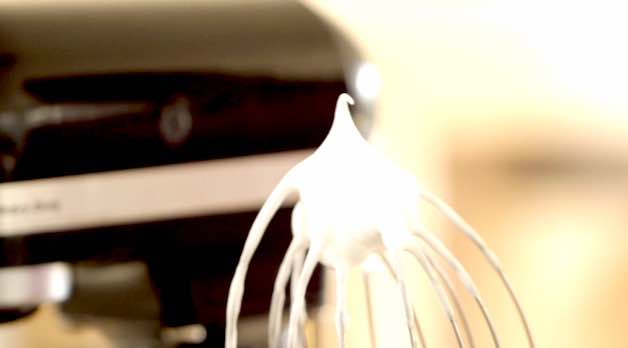 A stiff peak of whipped egg whites on a wire whisk attachment of a stand mixer