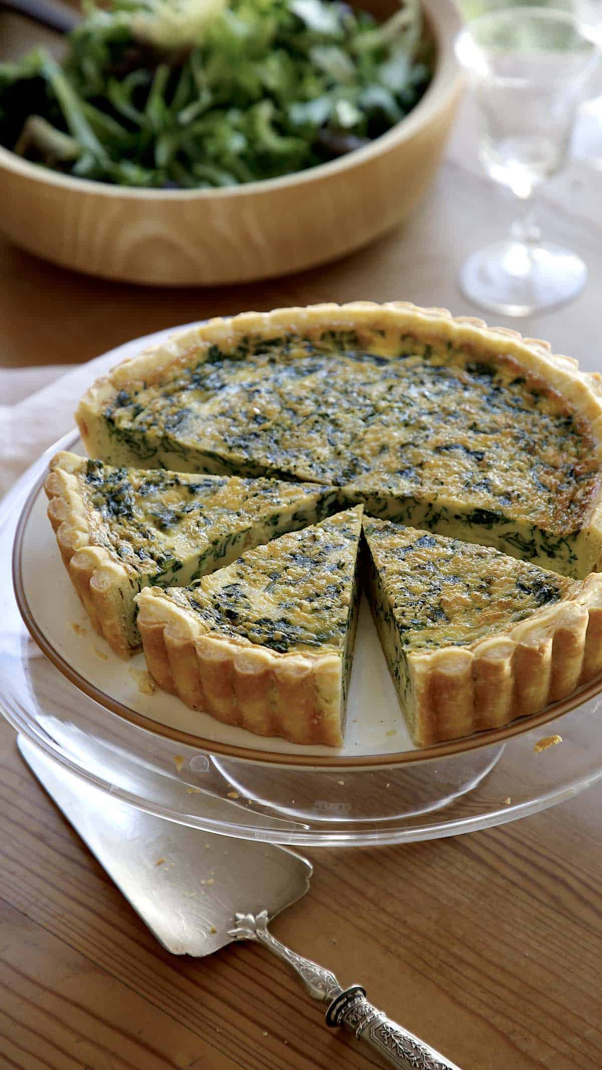 Spinach Quiche on a cake stand with slices cut and a salad bowl