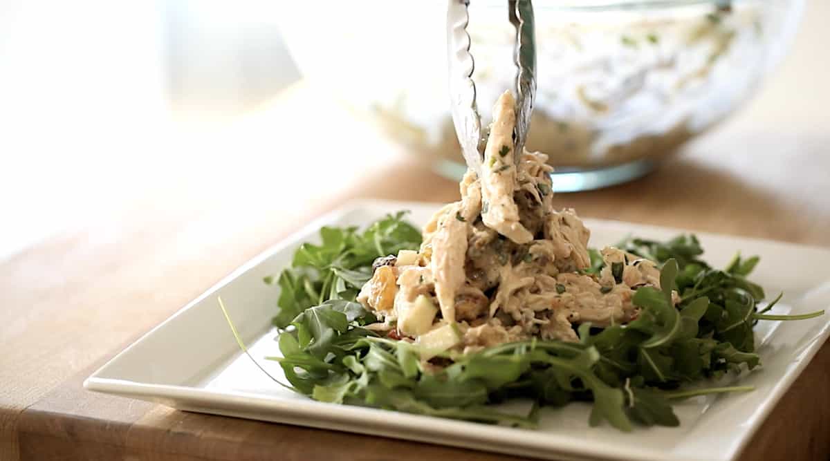a person adding chicken salad to a bed of arugula with tongs