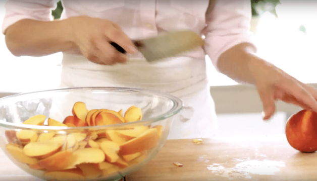 peaches being sliced on a wood board and added to a clear bowl