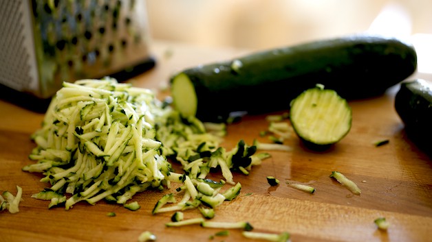 A wooden cutting board with zucchini being grated with a box grater