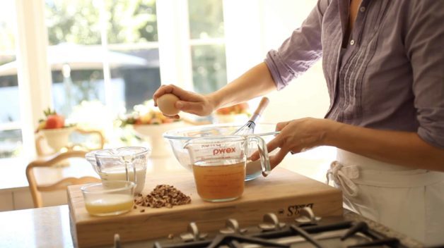 osoba rozklepávající vejce do průhledné misky na prkénku s odměřenými ostatními ingrediencemi