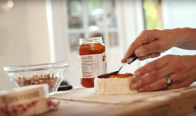 apricot jam being spread on top of a brie wheel with a spoon