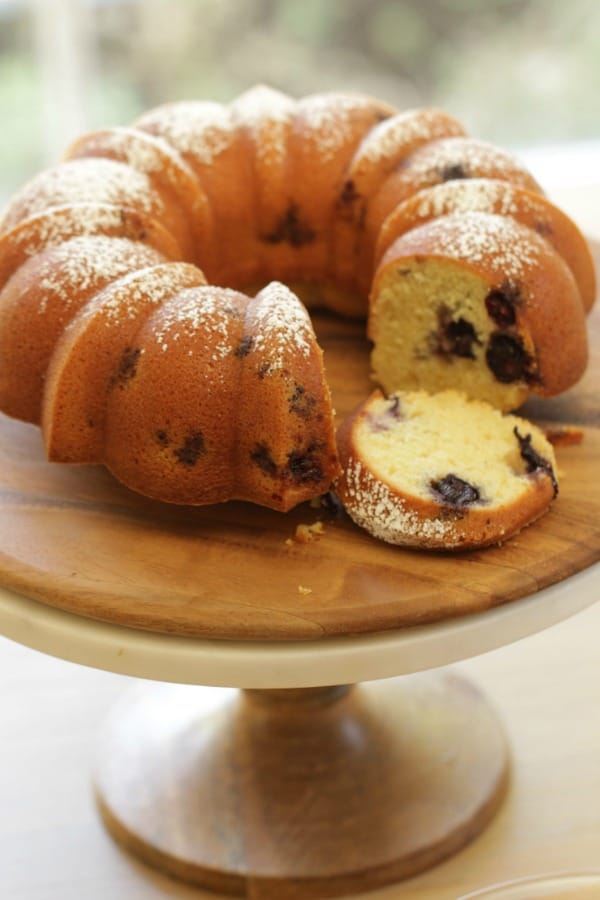 A verticle image of a Blueberry Lemon Bundt cake on a cake stand with a slice taken out