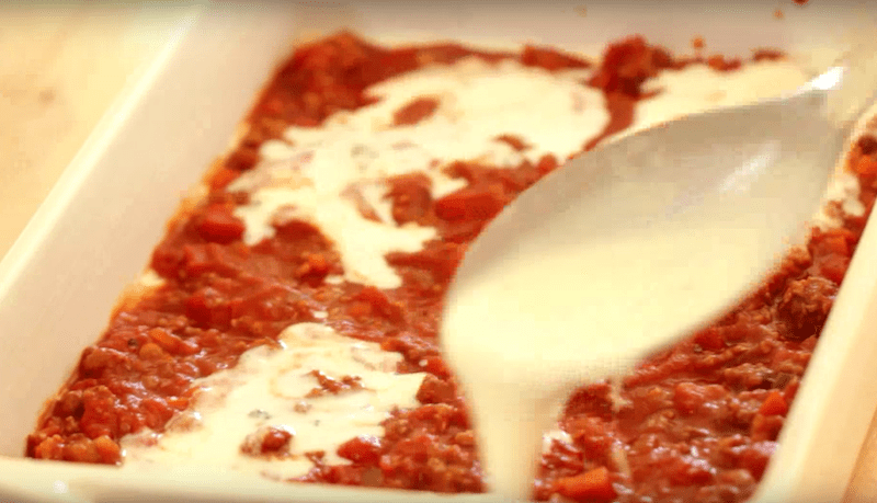 bechamel sauce being spooned on top of meat sauce in a lasagna pan