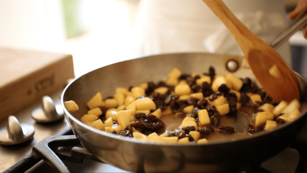 A wooden spoon stirring apples, raisins, and syrup in a skillet
