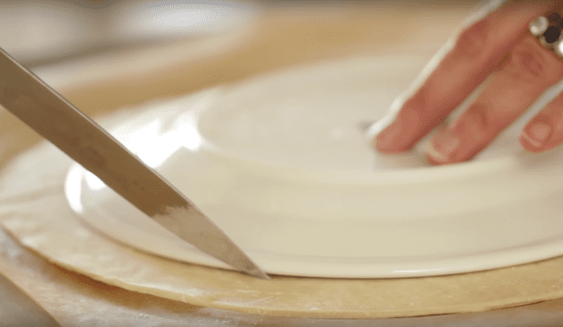 a knife cutting around a white plate on top of a sheet of puff pastry dough