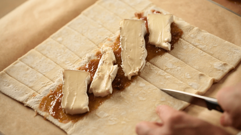 a person cutting slitsinto a sheet of puffed pastry with fig jam and cheese spread down the middle