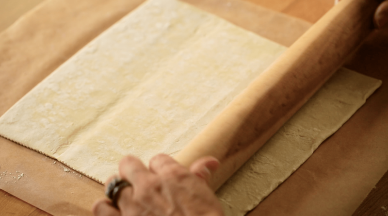 A person rolling puff pastry on a cutting board with a wooden rolling pin