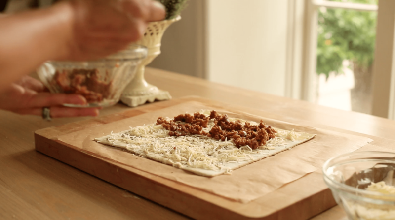 A sheet of puff pastry filled with grated cheese and cooked Italian Sausage on a table