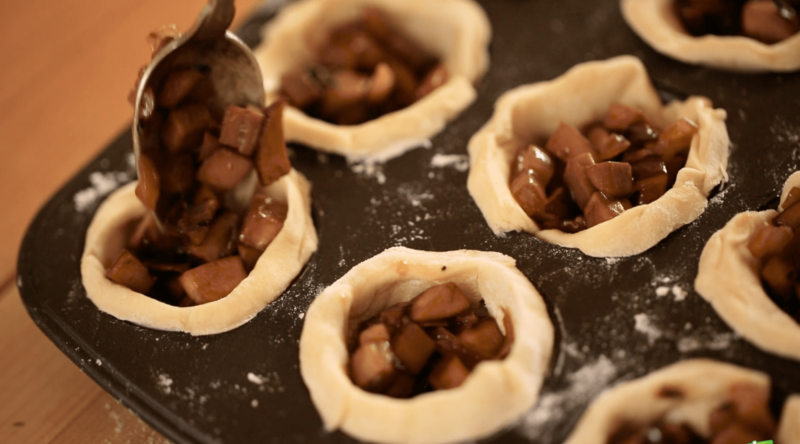 a spoon filling puff pastry tart shells in a muffin tin with a cooked mushroom mixture