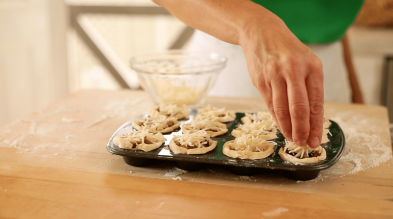 A person adding grated cheese to mini tartlets in a mini muffin tin