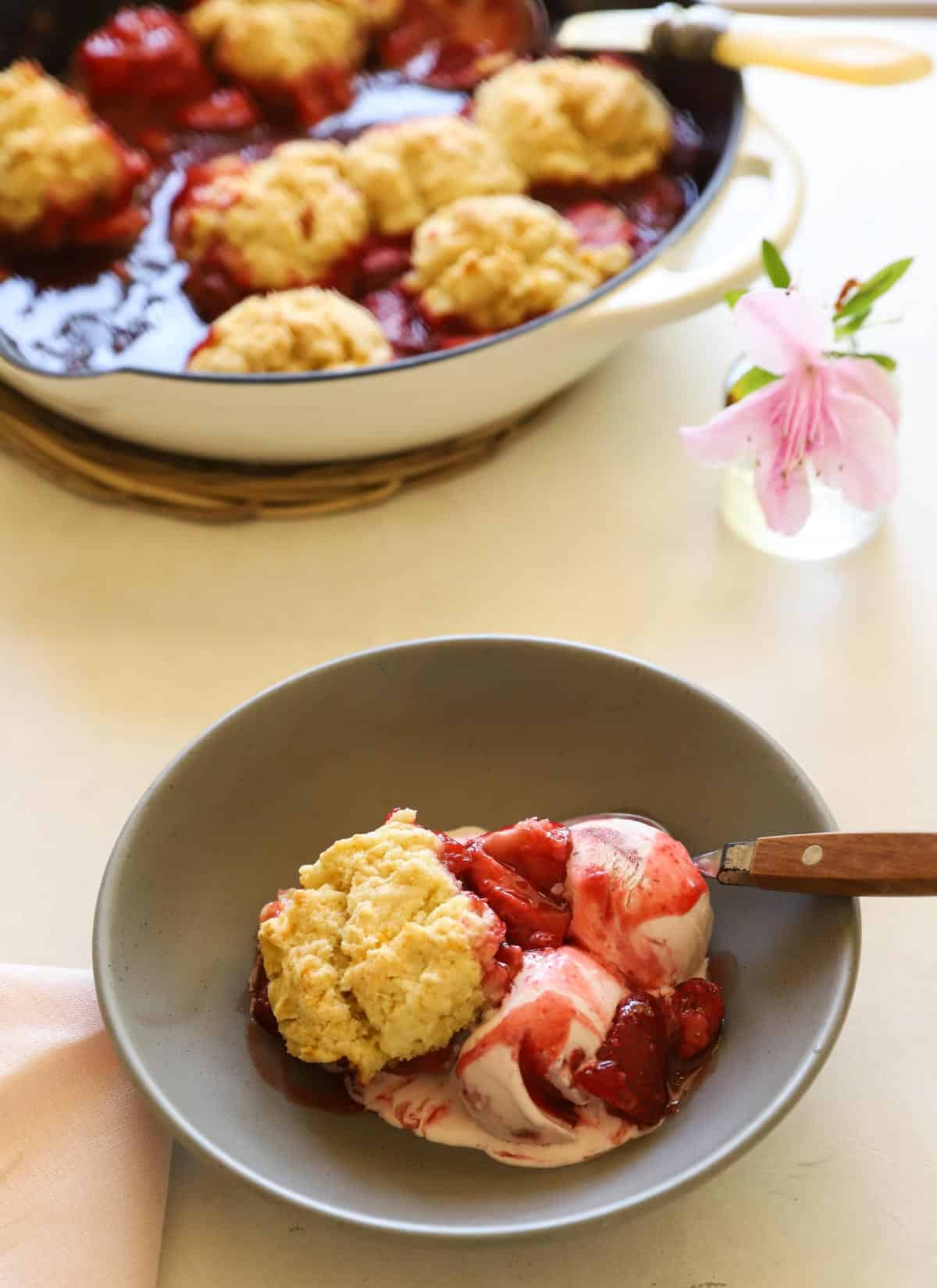 a cast-iron skillet filled with straberries and baked biscuits and a serving of strawberry cobbler with ice cream. 