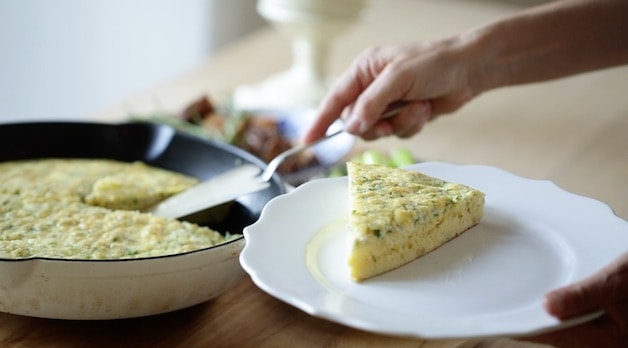 Slicing egg casserole from a skillet and placing a wedge on a white plate 