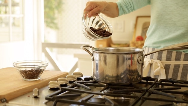 a person placing chocolate chips from a clear bowl into a pot on a cooktop