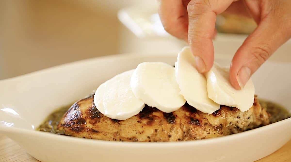 a person placing mozzarella cheese slices on a grilled chicken breast