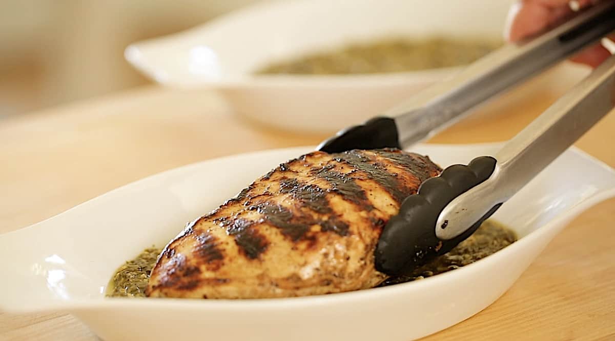 a person placing a grilled chicken breast into a gratin dish filled with pesto sauce