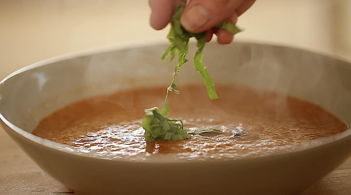 a person adding sliced basil to a roasted tomato soup