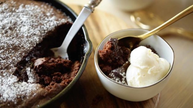Warm chocolate skillet cake in a cast-iron skillet with a bowl of cake scooped out of it topped with vanilla ice cream 