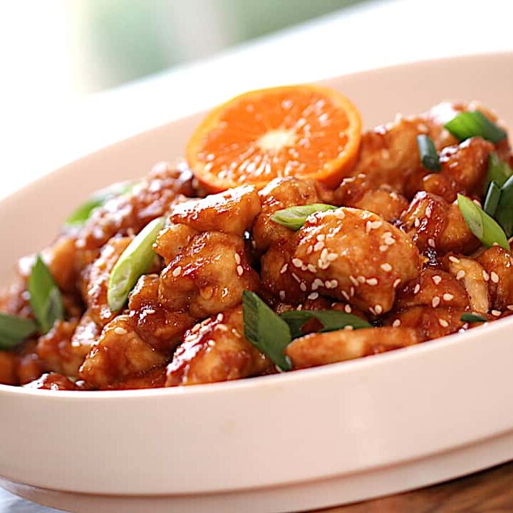 a finished plate of orange chicken garnished with green onions and a slice of orange