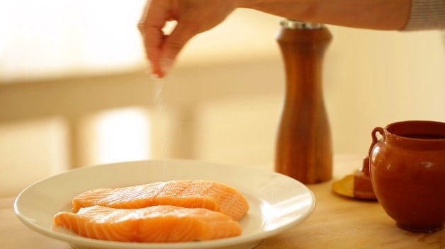 Salmon fillets being seasoned with salt on a white plate