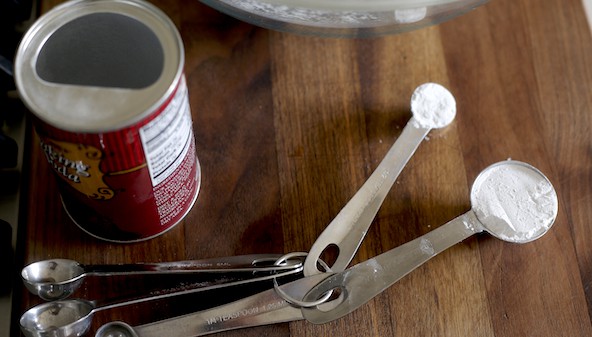 Measuring spoons on a dark cutting board filled with baking powder and baking soda