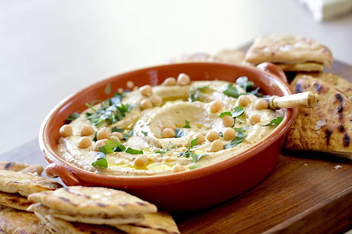 a bowl of homemade hummus garnished with parsley on a cutting board served with flatbreads