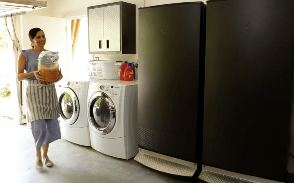 a person holding an aqua Fresca jar walking to a refrigerator and freezer in a garage