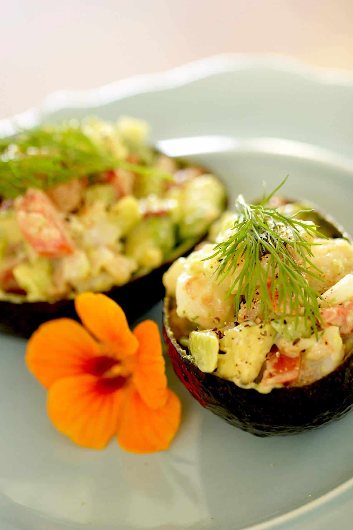 Two avocado shells stuffed with shrimp salad on a blue plate with a nasturtium