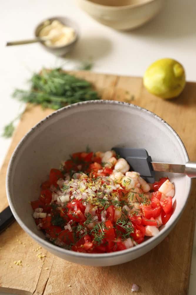 A bowl that includes shrimp, tomatoes, herbs, shallots and seasoning with a lemon and mayonnaise in the background.