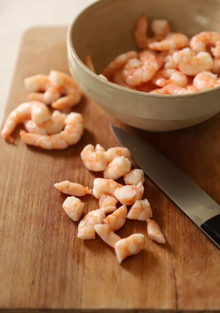 Diced shrimp on a cutting board with a bowl of cooked shrimp and a knife.