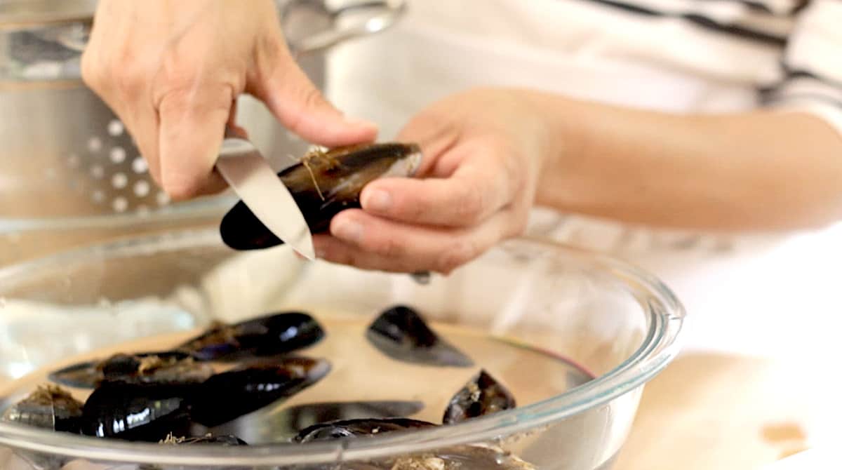 a woman holding a mussel over a bowl of water slicing off the beard
