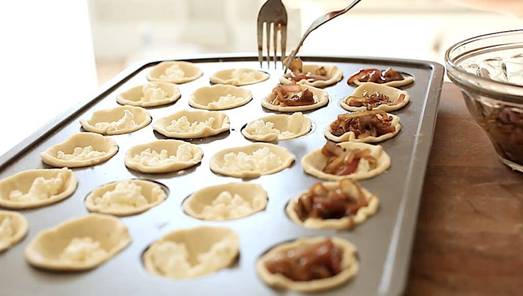 a person adding caramelized onions to the top of puff pastry and goat cheese in a mini muffin tin