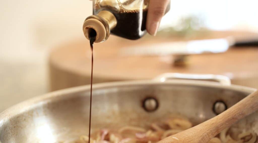 a person adding balsamic vinegar to onions cooking in a skillet
