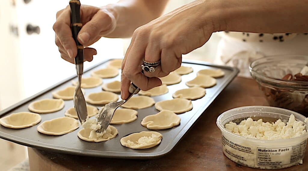 a person adding goat cheese crumbles into puff pastry crusts in a mini muffin tin
