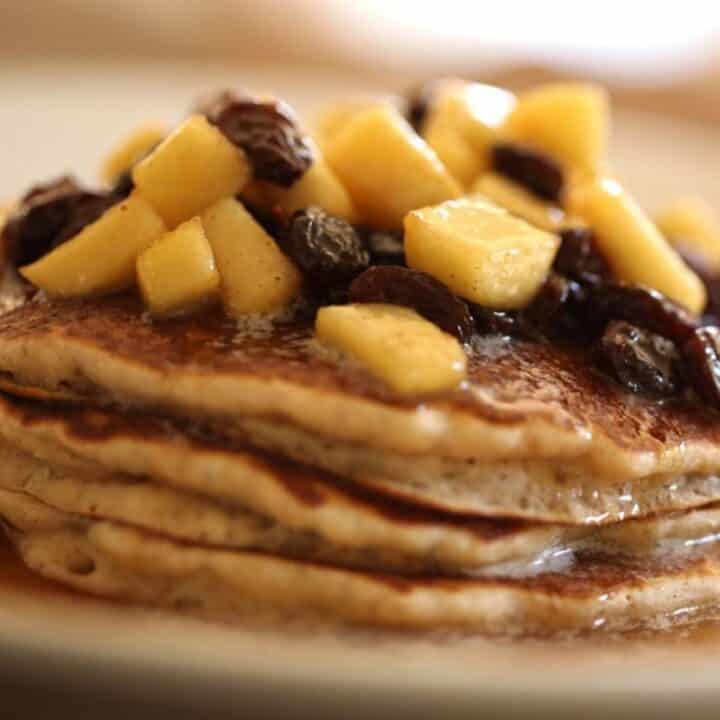 A stack of Apple Cider Pancakes with raisins and apples on top