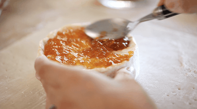 A close up of a spoon spreading apricot jam over a wheel of brie