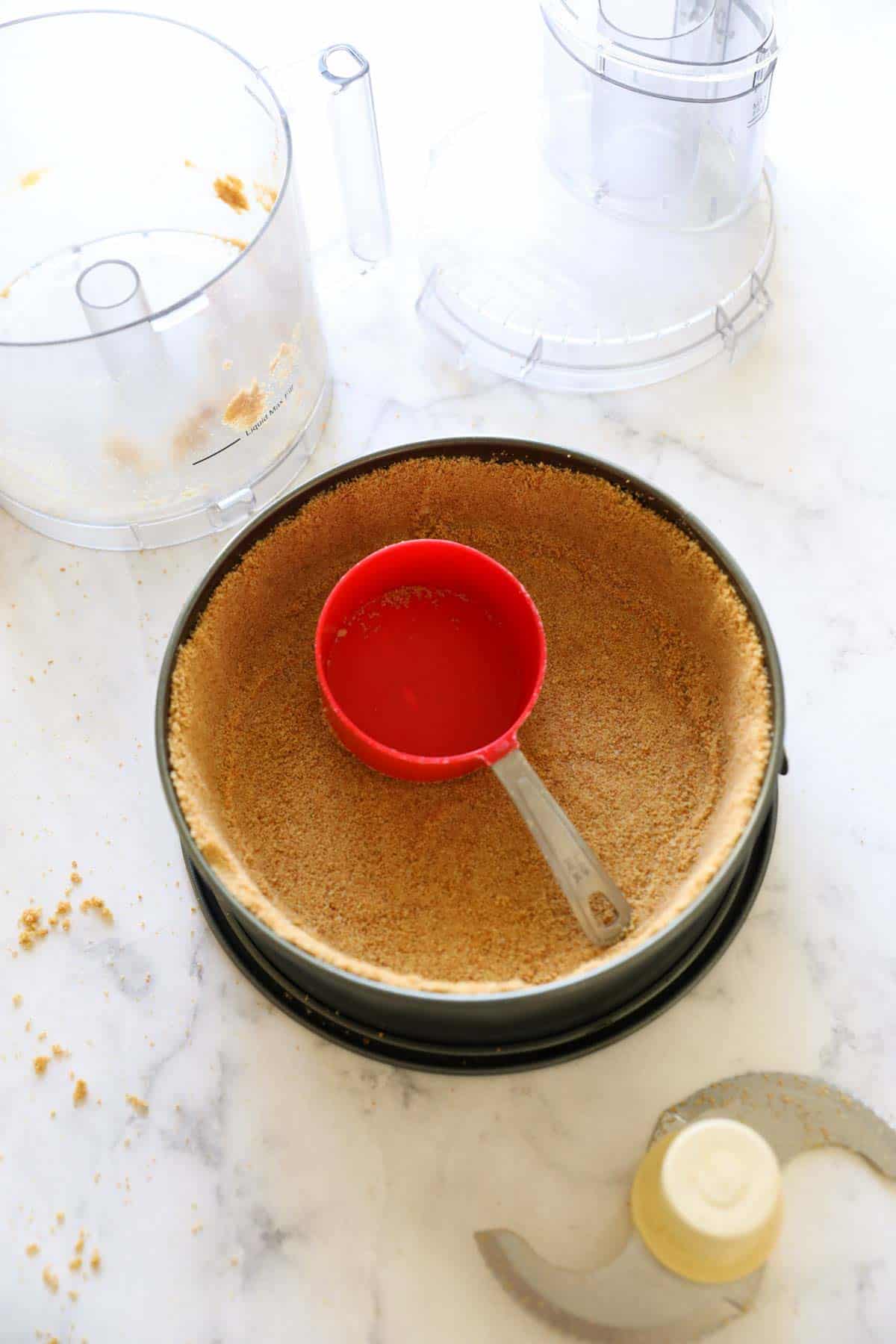 cheesecake pan with graham cracker crumbs being tamped down with a measuring cup