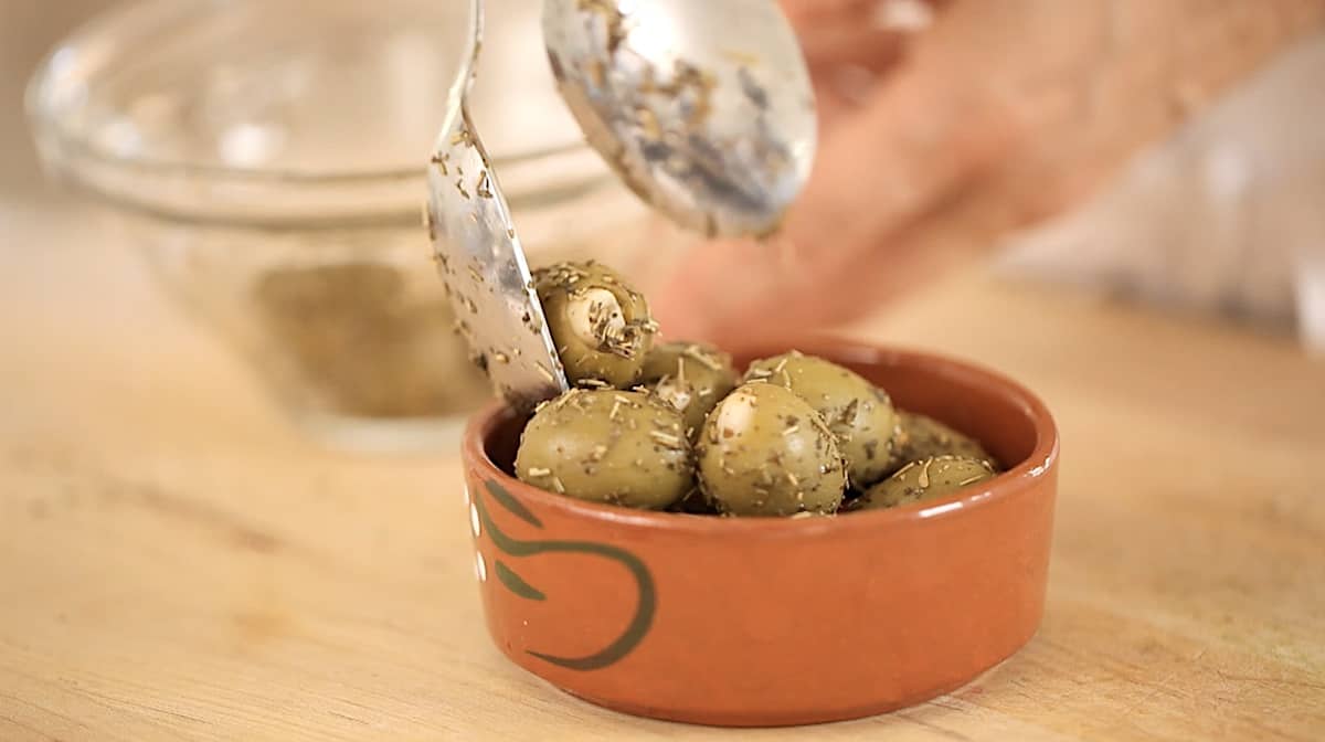 A person placing green olives with herbs de Provence in a bowl
