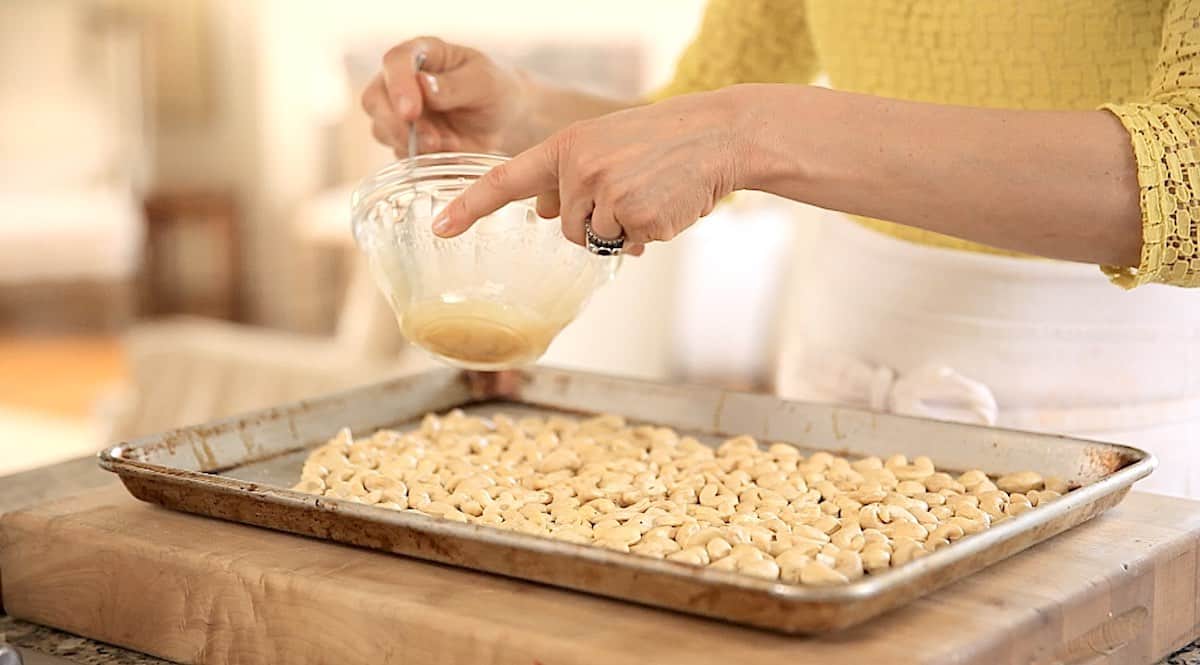 a sheet pan of cashews with a person about to add a honey glaze