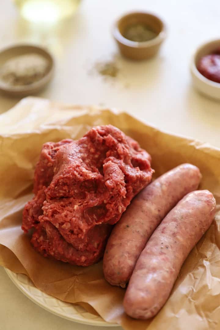 ground beef and Italian sausage links on a plate with seasoning in the background