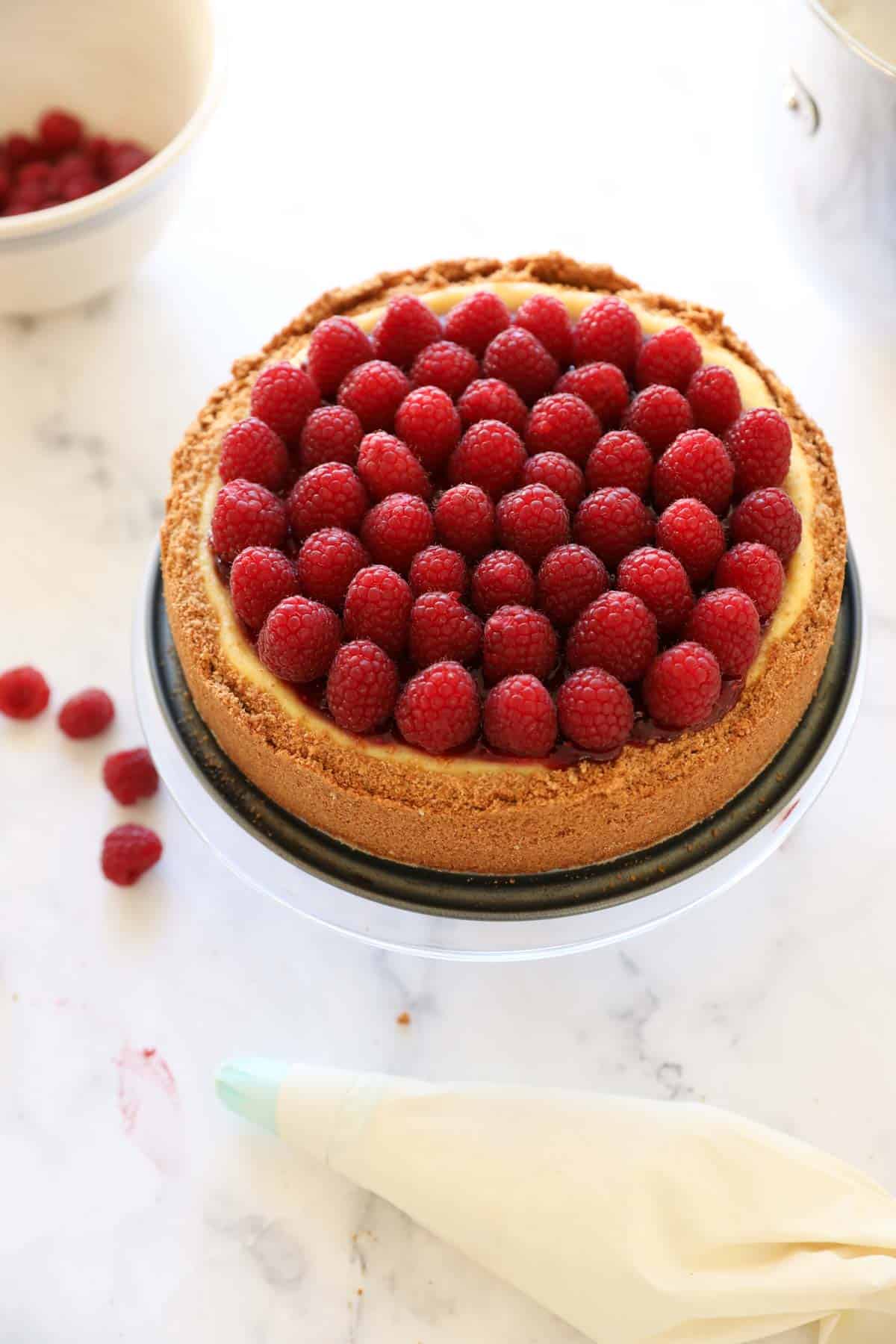 A full raspberry cheesecake decorated with raspberry jam and fresh raspberries on a cake stand