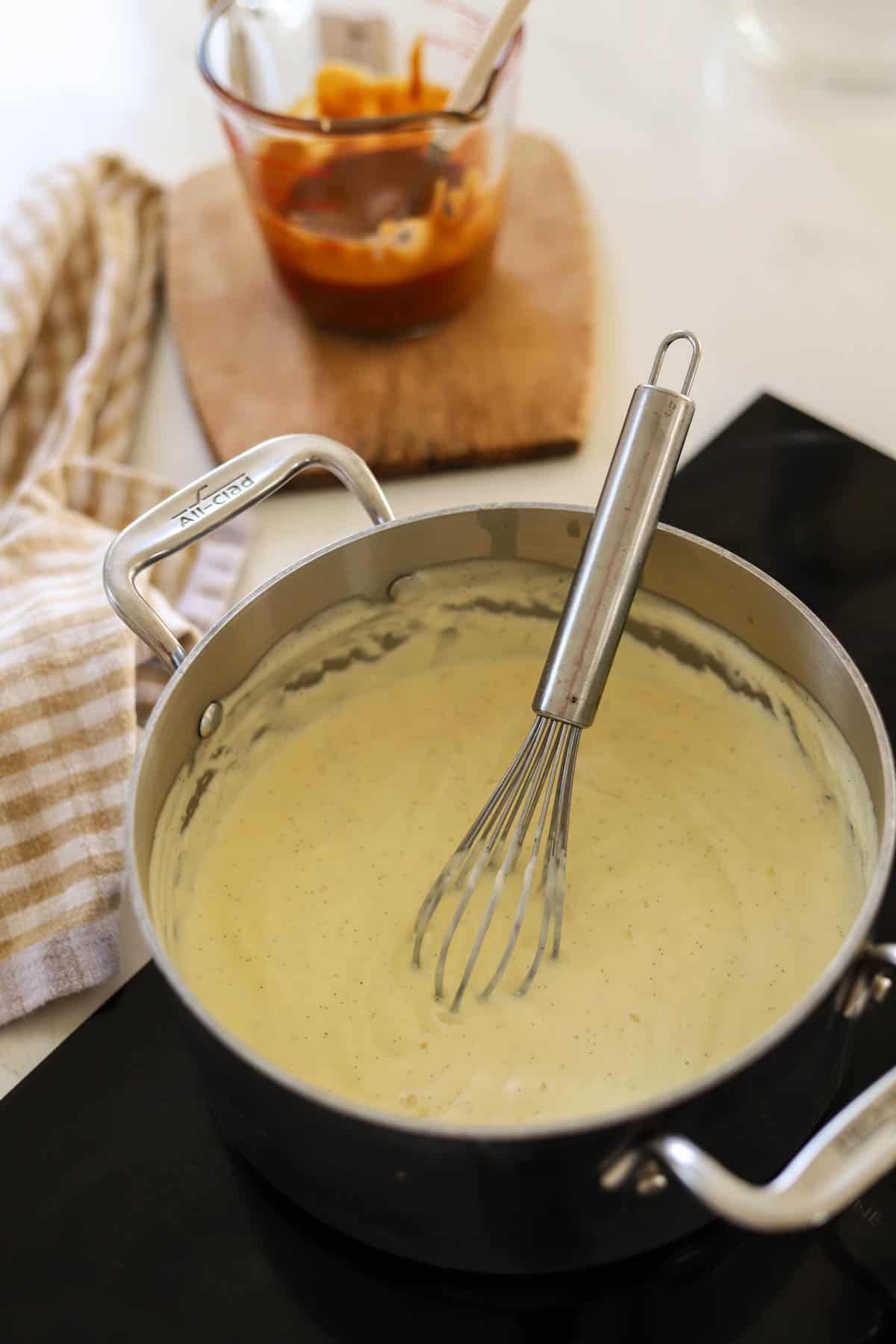 A vanilla pudding on the stove with whisk, with salted caramel in a pitcher in the background.