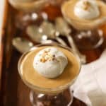 Salted Caramel Puddings in glass dishes topped with whipped cream and toffee bits on a wooden tray.