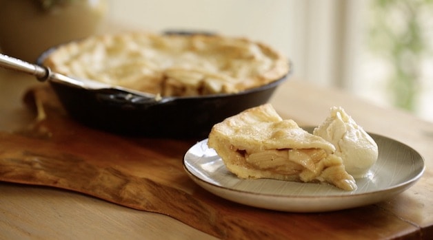 A slice of apple pie with a scoop of vanilla ice cream in the foreground with a whole apple pie in the background