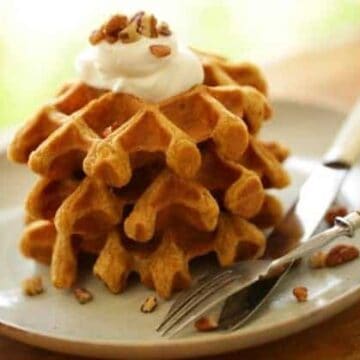 Pumpkin Spice Waffles stacked on a plate topped with whipped cream and chopped pecans