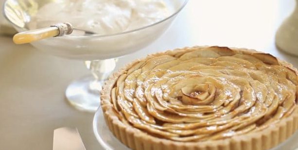 French Apple Tart on a clear cake stand with whipped cream in the background